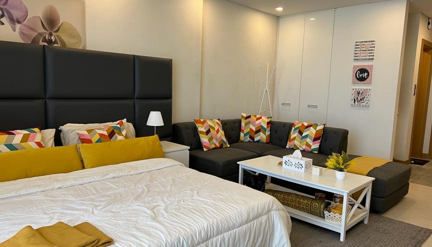 A modern bedroom with a large bed, gray sectional sofa with colorful cushions, white coffee table, and wall art, featuring neutral walls and bright yellow accents.