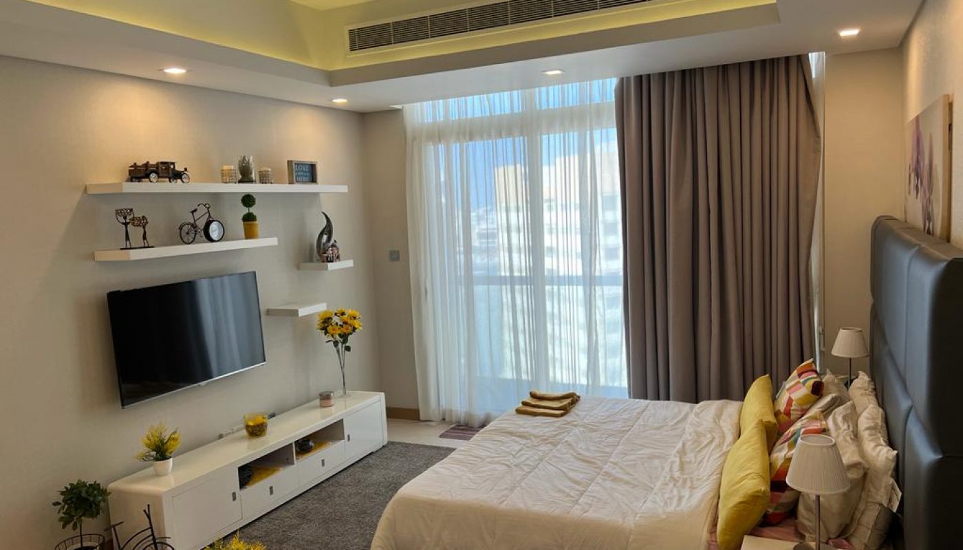 Modern bedroom with a large window, white furniture, wall-mounted TV, decorative shelves, and a neatly made bed with colorful pillows and folded towels.