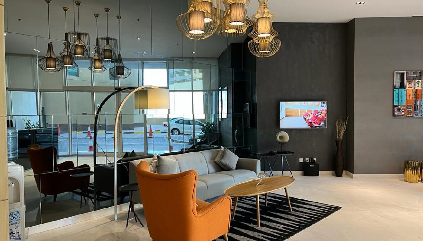 Modern lobby with a gray sofa, orange armchair, round wooden tables, striped rug, large mirrors, wall-mounted TV, and decorative hanging lights.