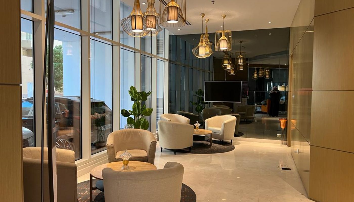 Modern lounge area with beige armchairs, round tables, pendant lights, large windows, indoor plants, and a flat-screen TV in the background.