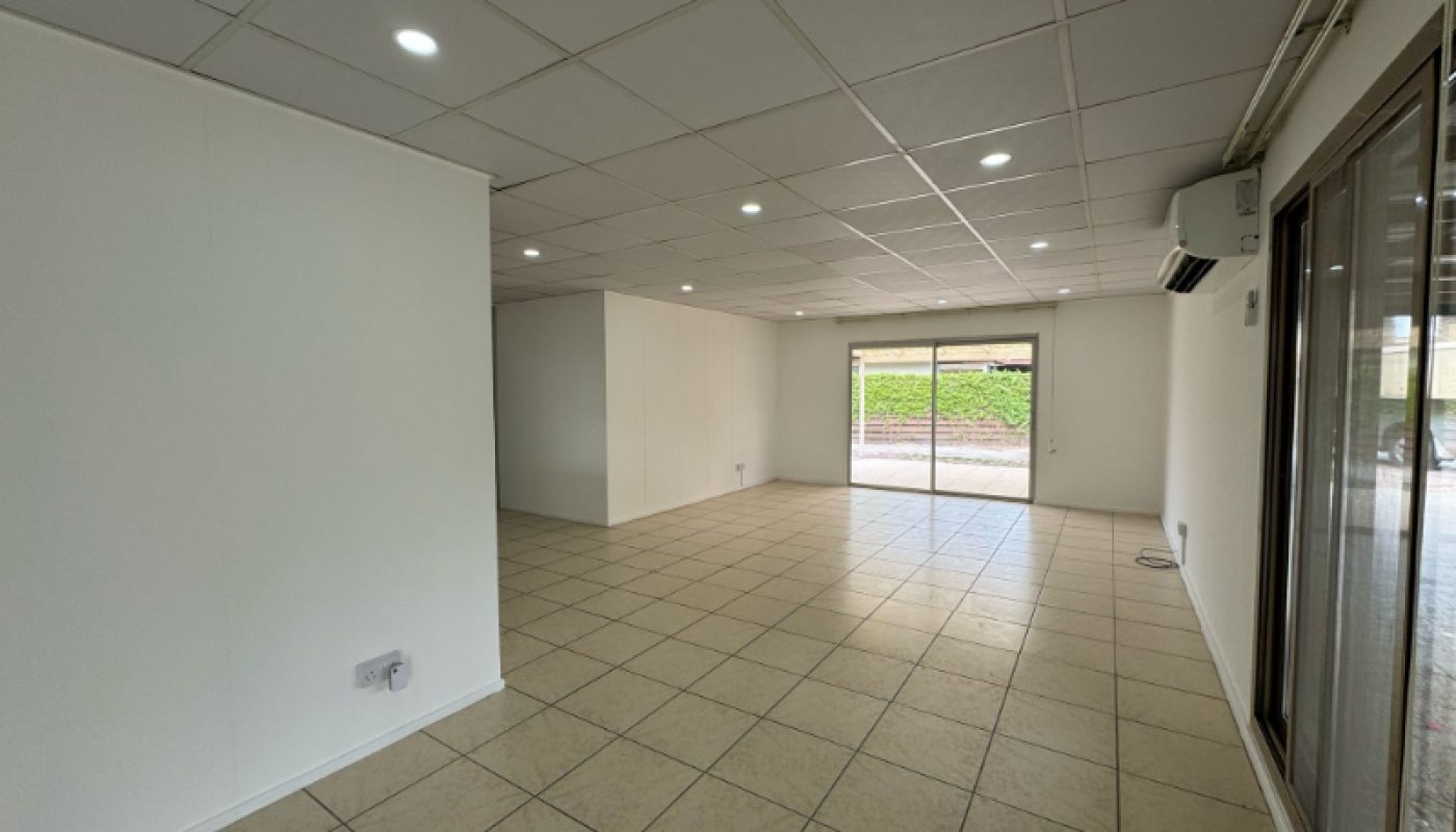 Empty room with tiled floor, white walls, and a grid-patterned ceiling with recessed lights. Large windows and glass door provide natural light and outdoor view.