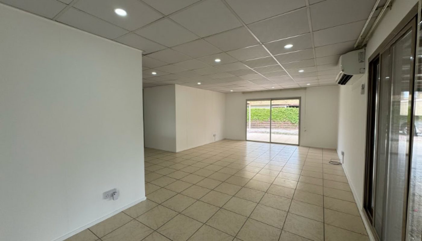 Empty room with tiled floor, white walls, and a grid-patterned ceiling with recessed lights. Large windows and glass door provide natural light and outdoor view.