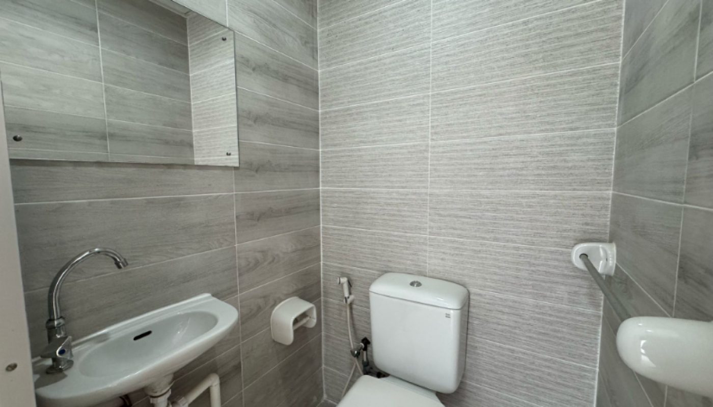 A small bathroom with grey tiled walls, a wall-mounted sink with exposed pipes, a toilet, a mirror, a toilet paper holder, and a grab bar.