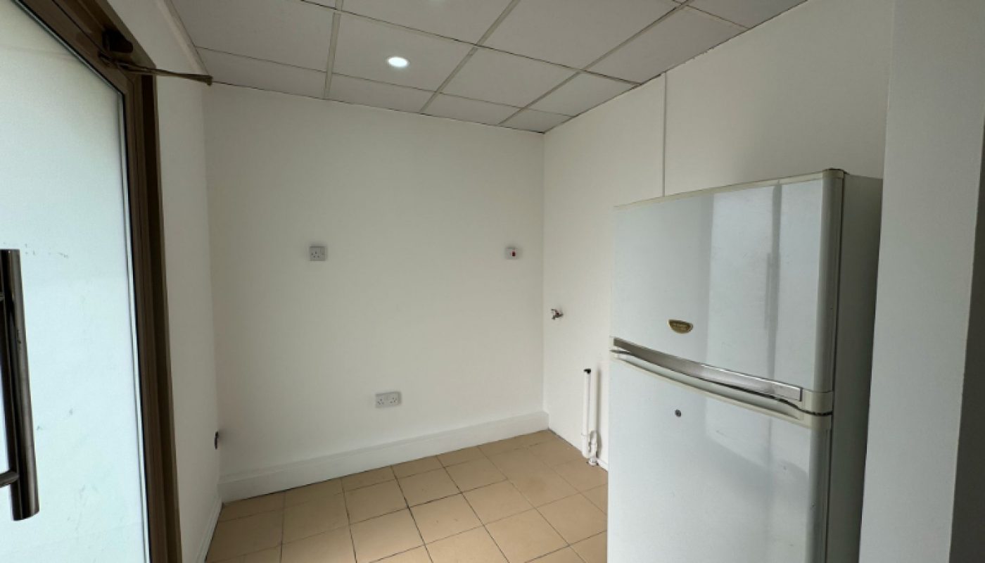 Small, unfurnished room with tiled floor, white walls, ceiling lights, a large windowed door, and a white fridge positioned in the corner.