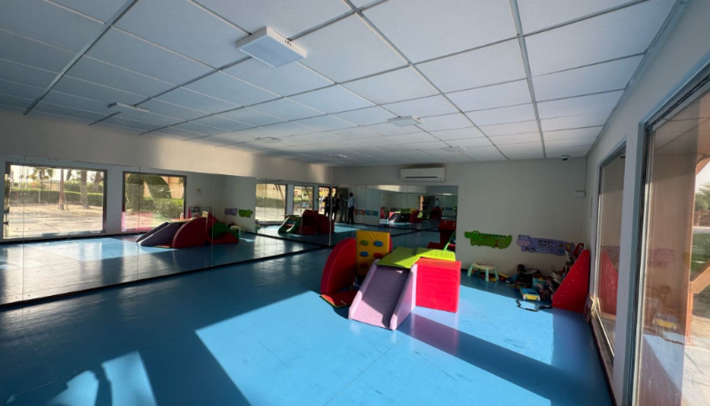 Room with blue padded floor, wall mirrors, large windows, foam play structures, and children’s toys scattered around.