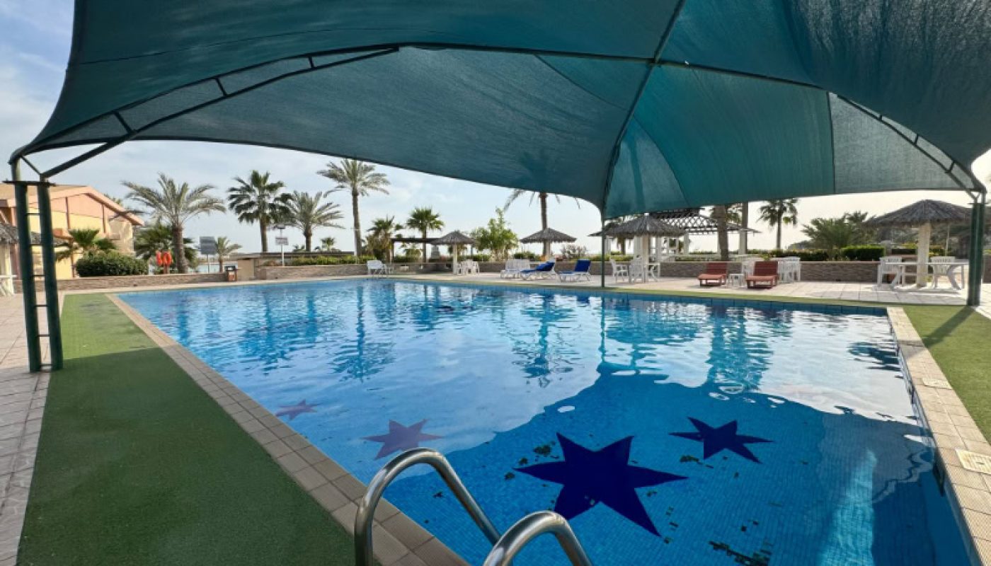 Outdoor swimming pool with blue water and star designs, partially covered by a fabric canopy, surrounded by lounge chairs and palm trees.