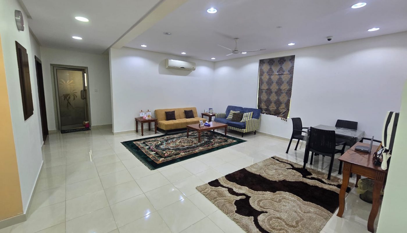 Spacious living room with tiled floor, sofas, coffee table, two area rugs, dining table with four chairs, wall-mounted AC, and ceiling fan.