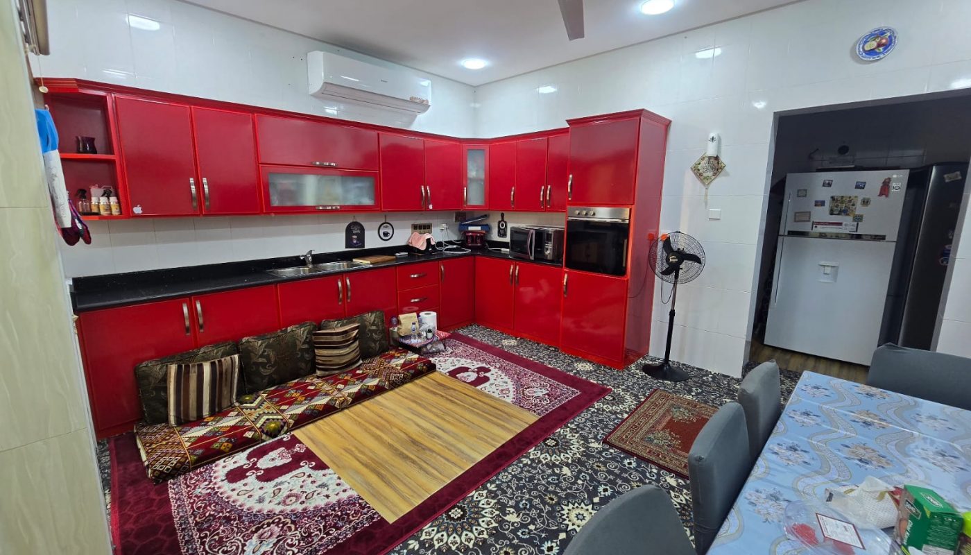 A modern kitchen with red cabinets, a wall-mounted AC, ceiling fan, patterned rugs, floor cushions around a low table, and a dining table with chairs. A refrigerator is visible in an adjacent room.