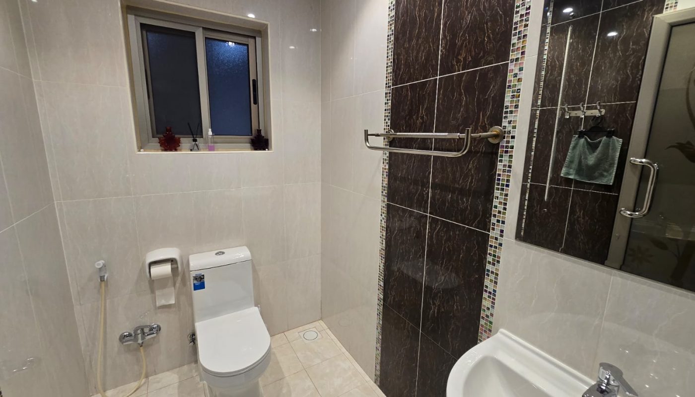 A modern bathroom with beige tiled walls, a white toilet, a towel rack, a wall mirror above a sink, and a window with frosted glass.