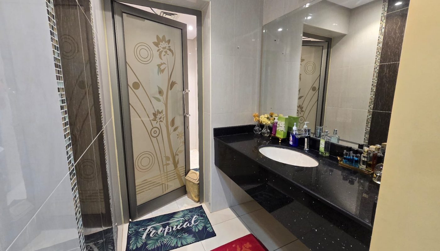 Modern bathroom with a black countertop, sink, assorted toiletries, a large mirror, decorative door, and two floor mats on white tile flooring.