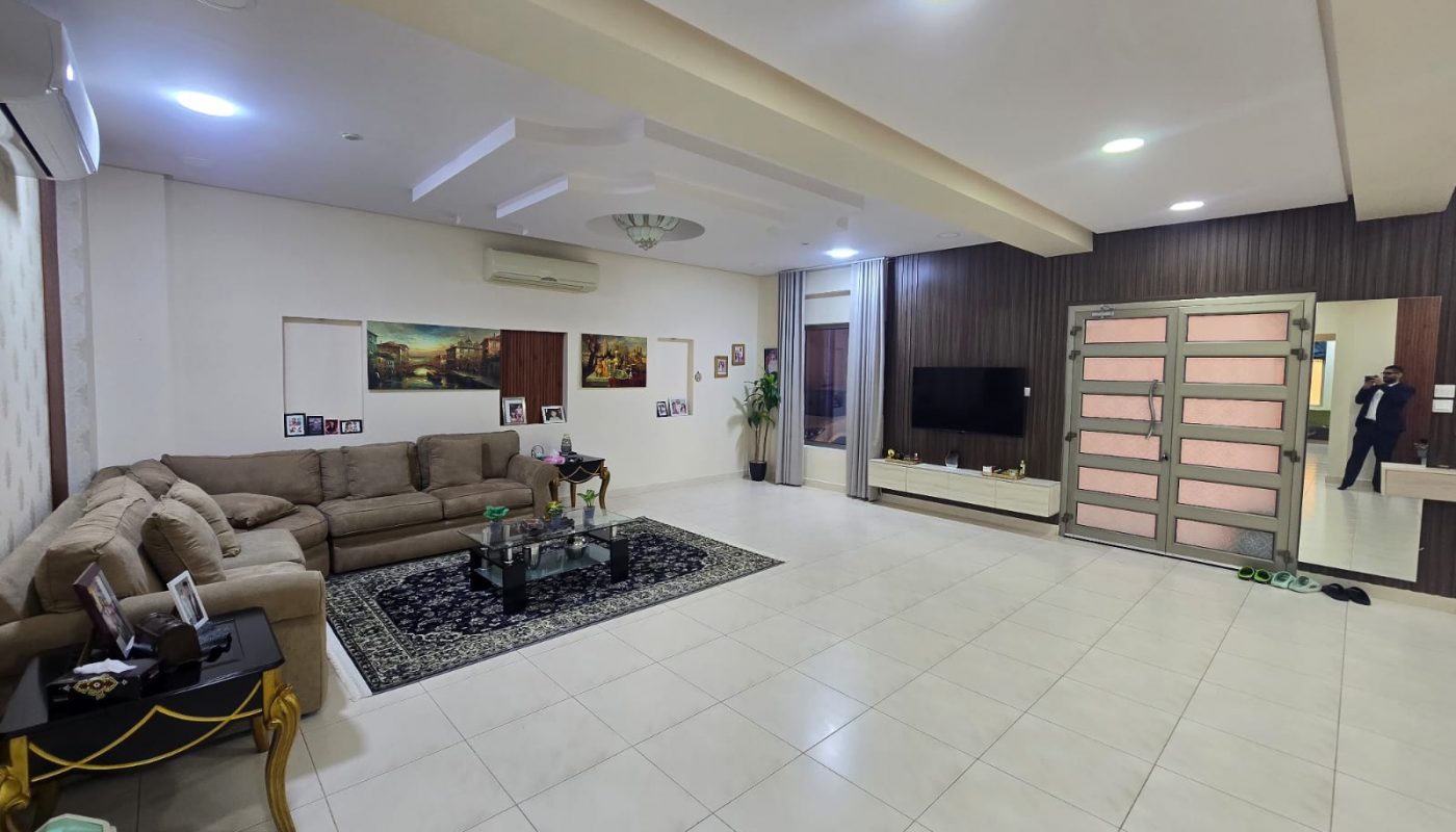 Spacious living room with beige sectional sofa, patterned rug, wall-mounted TV, paintings on the wall, and pink double doors; a person is visible near the entrance.