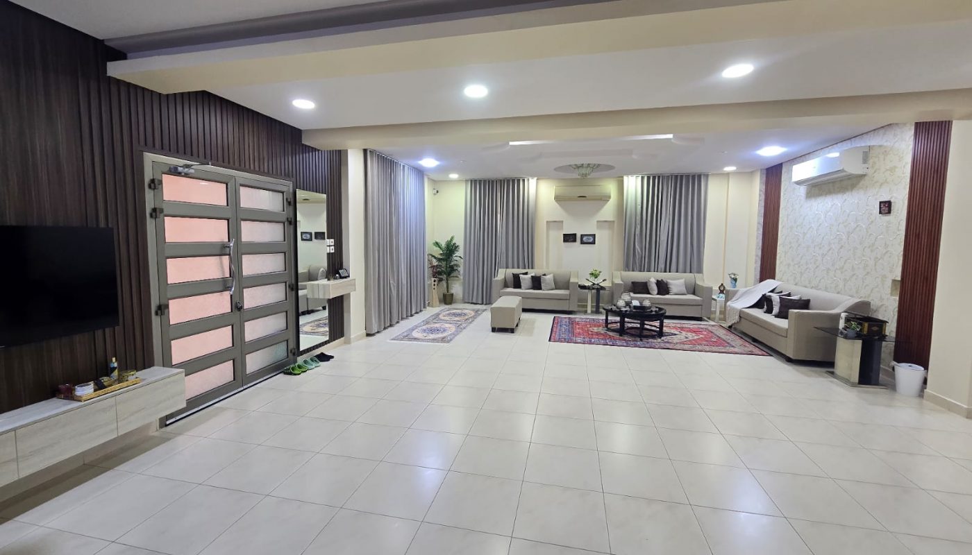 Spacious living room with tiled floor, light-colored sofas, area rugs, a round coffee table, wall-mounted TV, large windows with curtains, and modern ceiling lights.