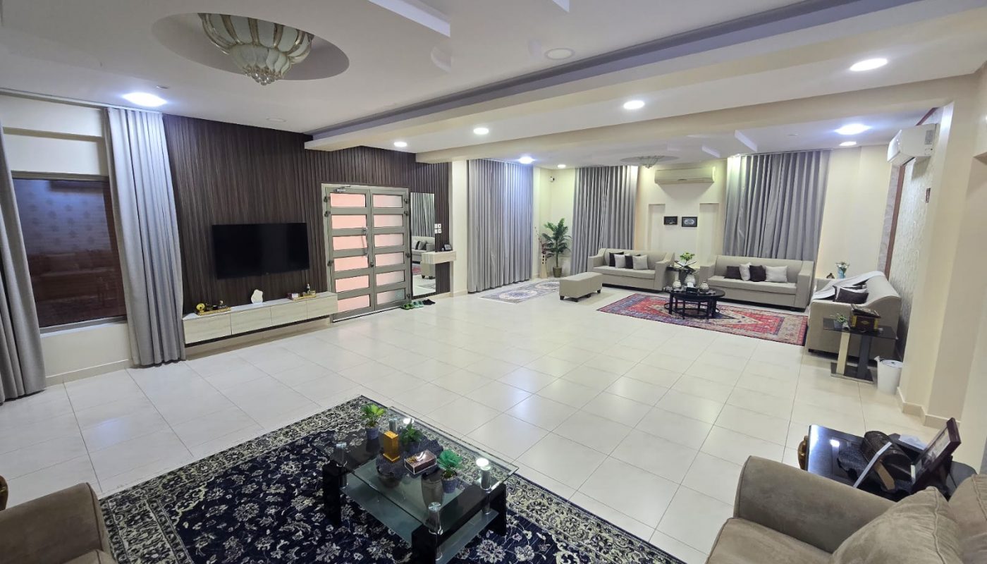 Spacious modern living room with tiled floor, multiple seating areas, wall-mounted TV, large rug, glass coffee table, decorative plants, and ceiling lights.