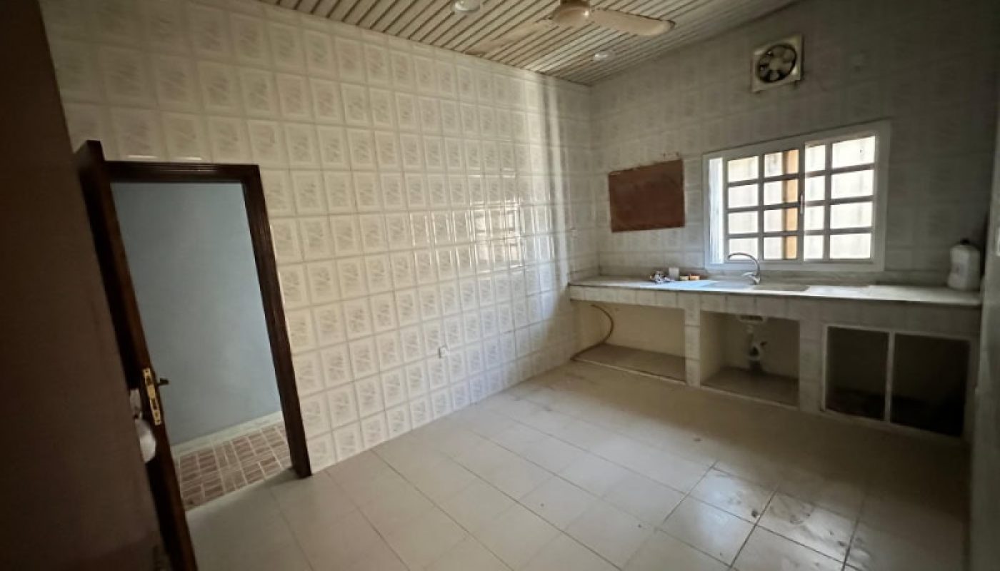 Empty tiled kitchen with a ceiling fan, open shelves under the countertop, a window, and a sink; an open door leads to another room.