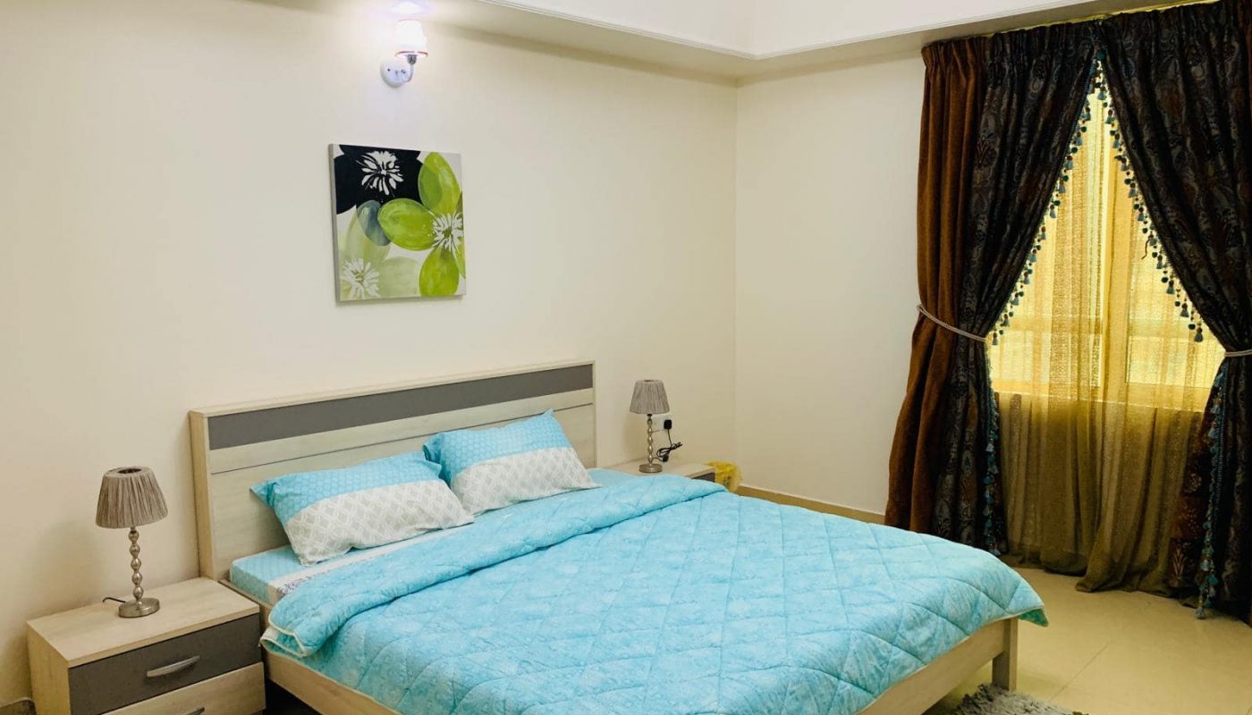 A bedroom with a double bed covered in a blue quilt, two bedside tables with lamps, a window with brown curtains, and a floral painting above the bed.