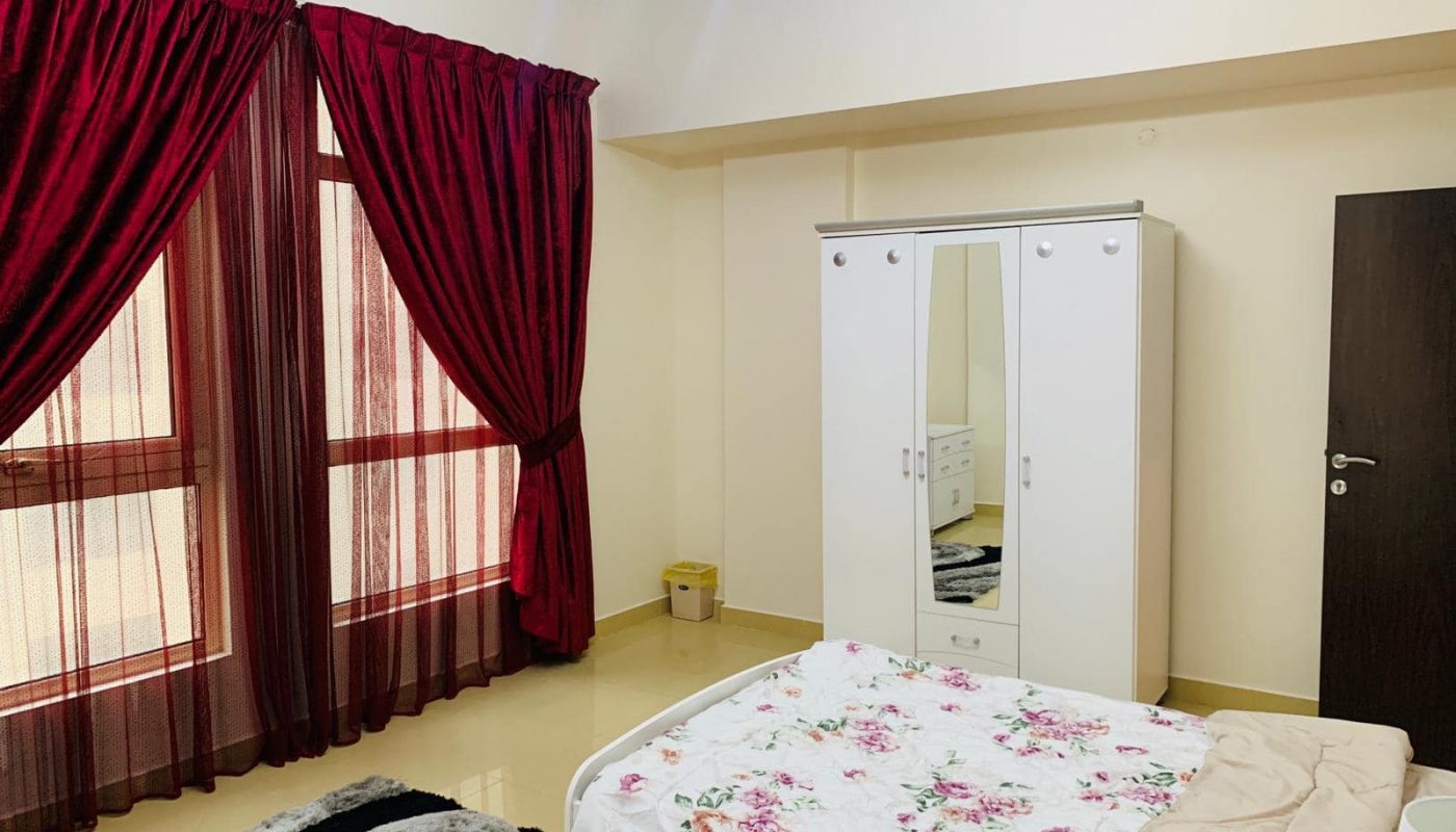 A bedroom with two beds featuring floral bedding, a white wardrobe with a mirror, dark red curtains, a beige wall, and a chandelier on the ceiling.