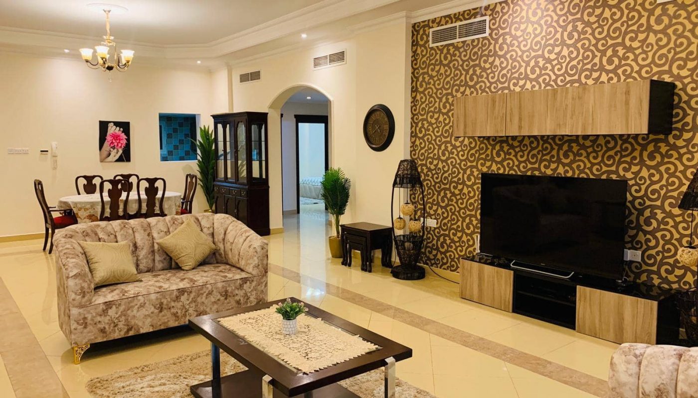 Spacious living room with patterned accent wall, TV, beige sofas, coffee table, and dining area in the background with a chandelier overhead.