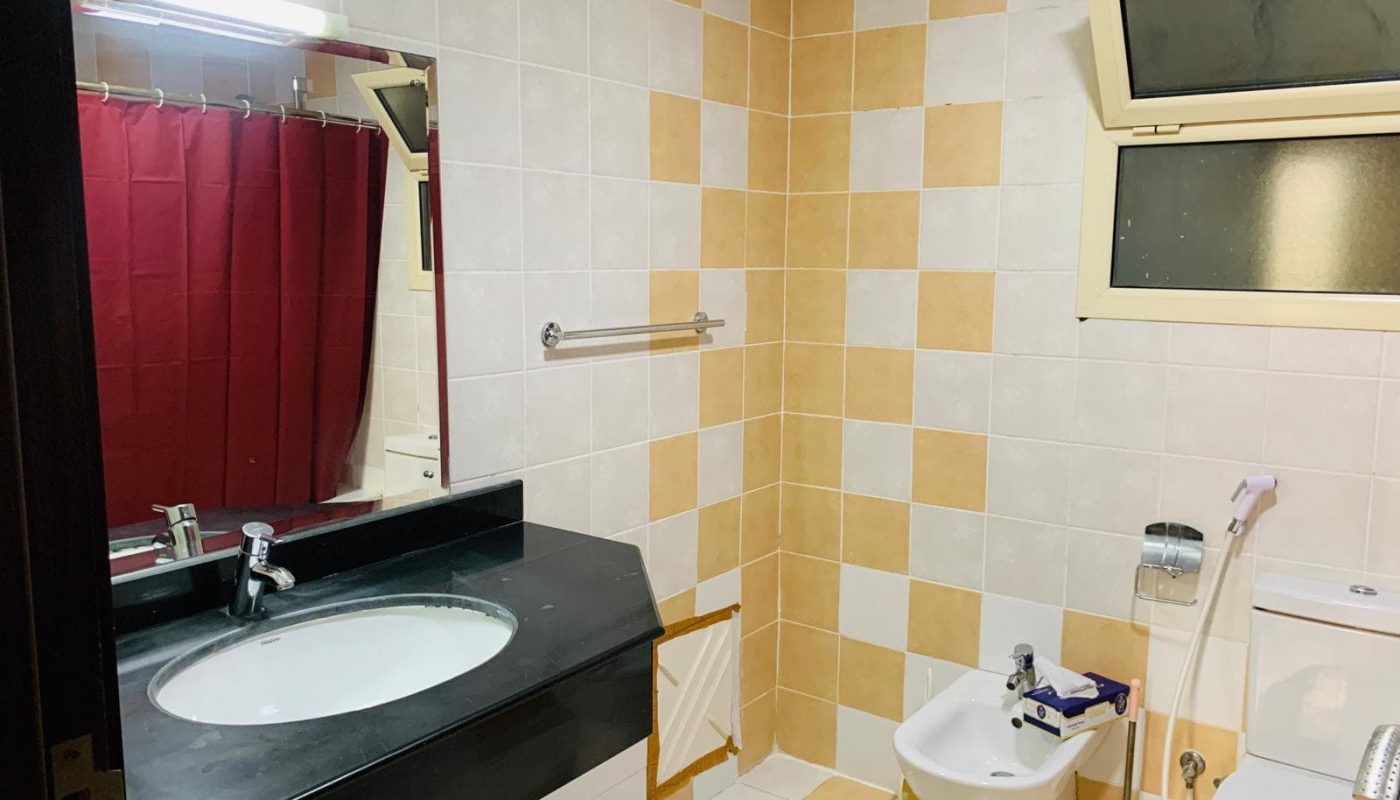 A bathroom with a sink, mirror, bidet, toilet, towel rack, red shower curtain, and a window, featuring yellow and white tiled walls and floor.