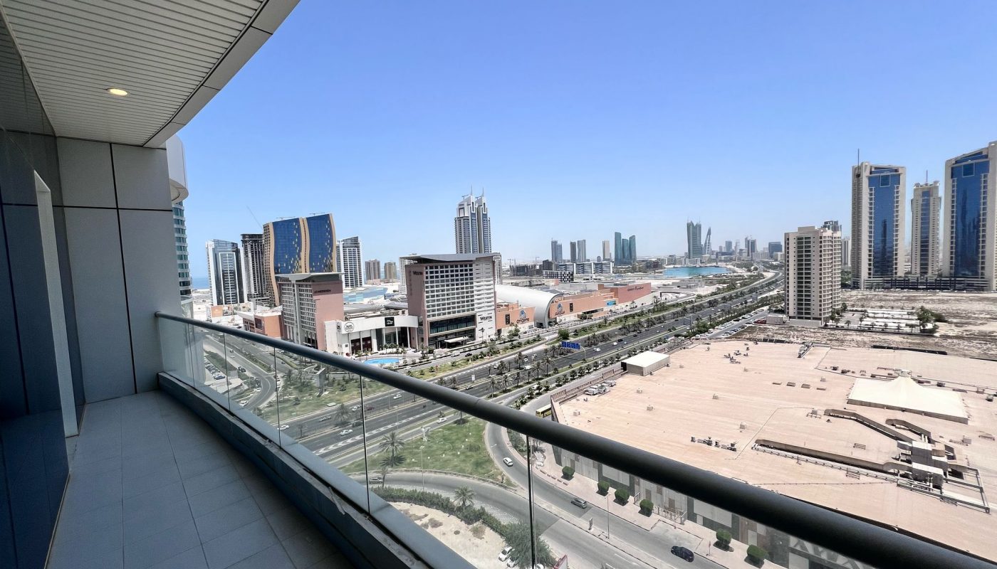 City skyline view from a high-rise balcony with glass railing, overlooking modern buildings, a mall, and main roads under a clear blue sky.