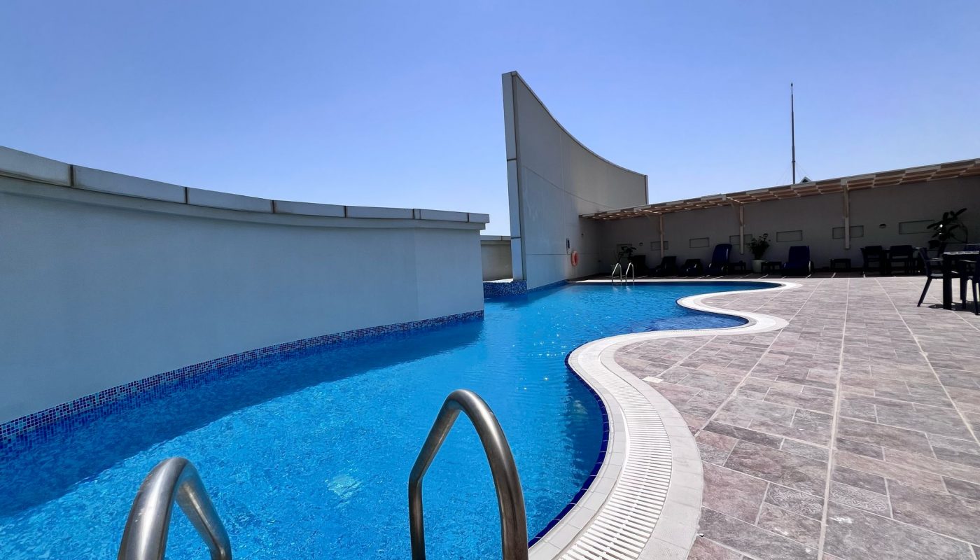 A rooftop swimming pool with clear blue water, metal handrails, and poolside lounge chairs under a shaded area on a sunny day.
