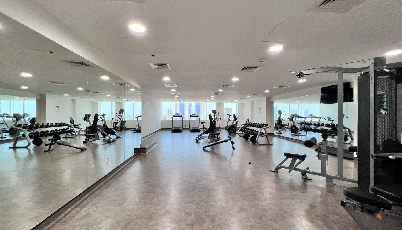 Spacious gym room with exercise equipment, free weights, benches, and mirrored walls; large windows provide natural light and a city view.
