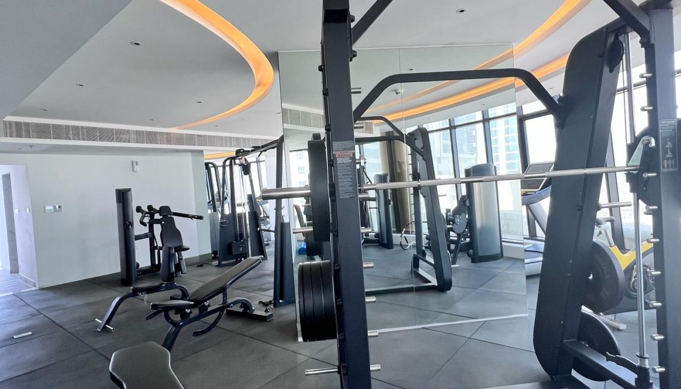Modern gym with various exercise machines, a weight bench, free weights, and large windows allowing natural light into the space.