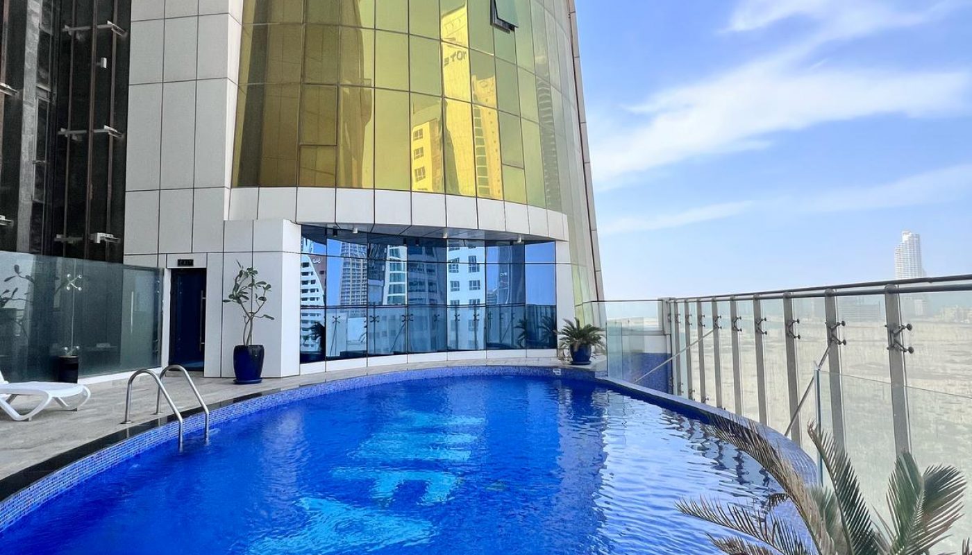 A rooftop swimming pool with a blue tiled bottom and silver railings, situated next to a modern building with golden windows.