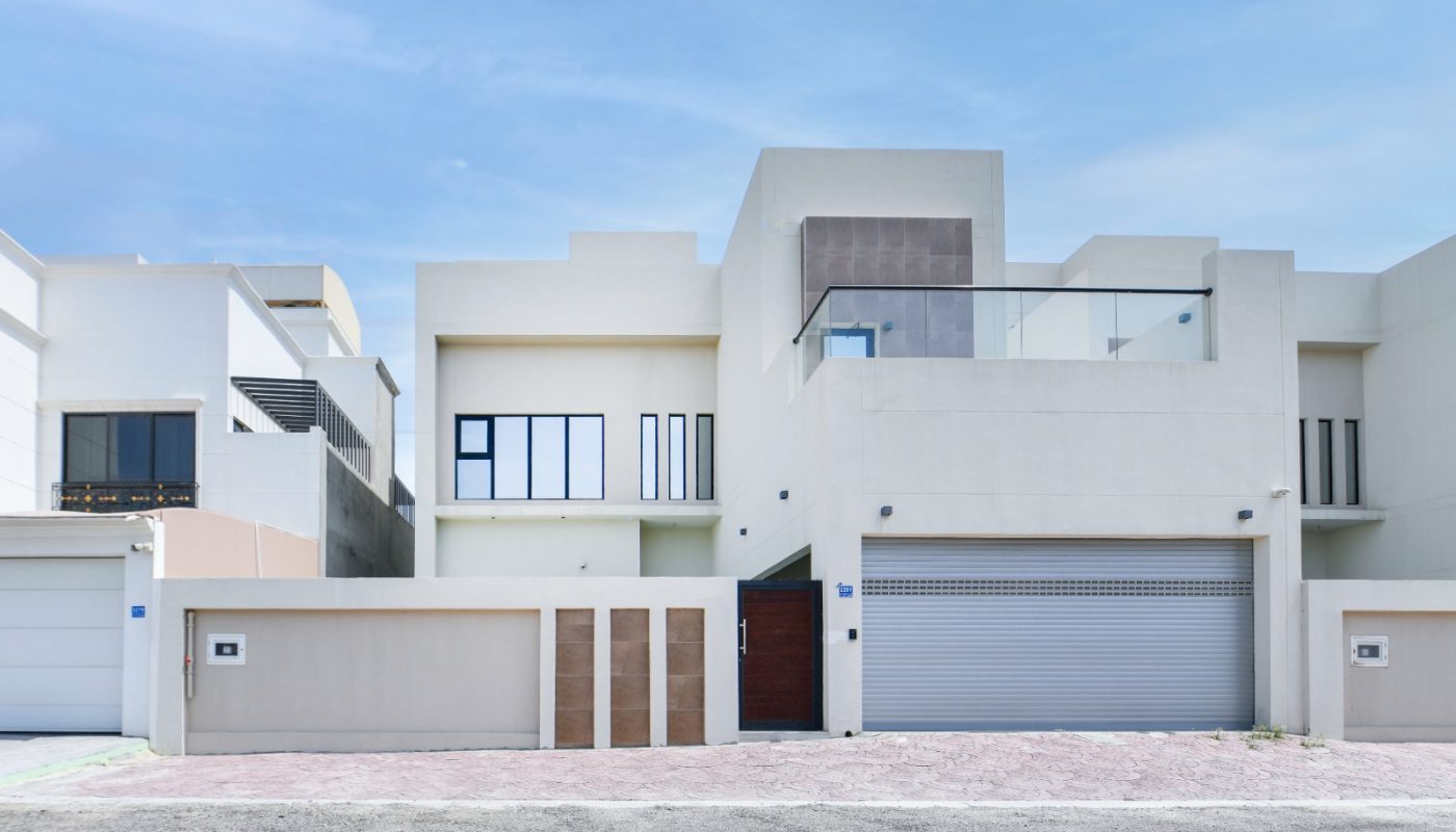 A modern, two-story white house with geometric design elements, large windows, a balcony, and a closed garage door, set against a clear blue sky.