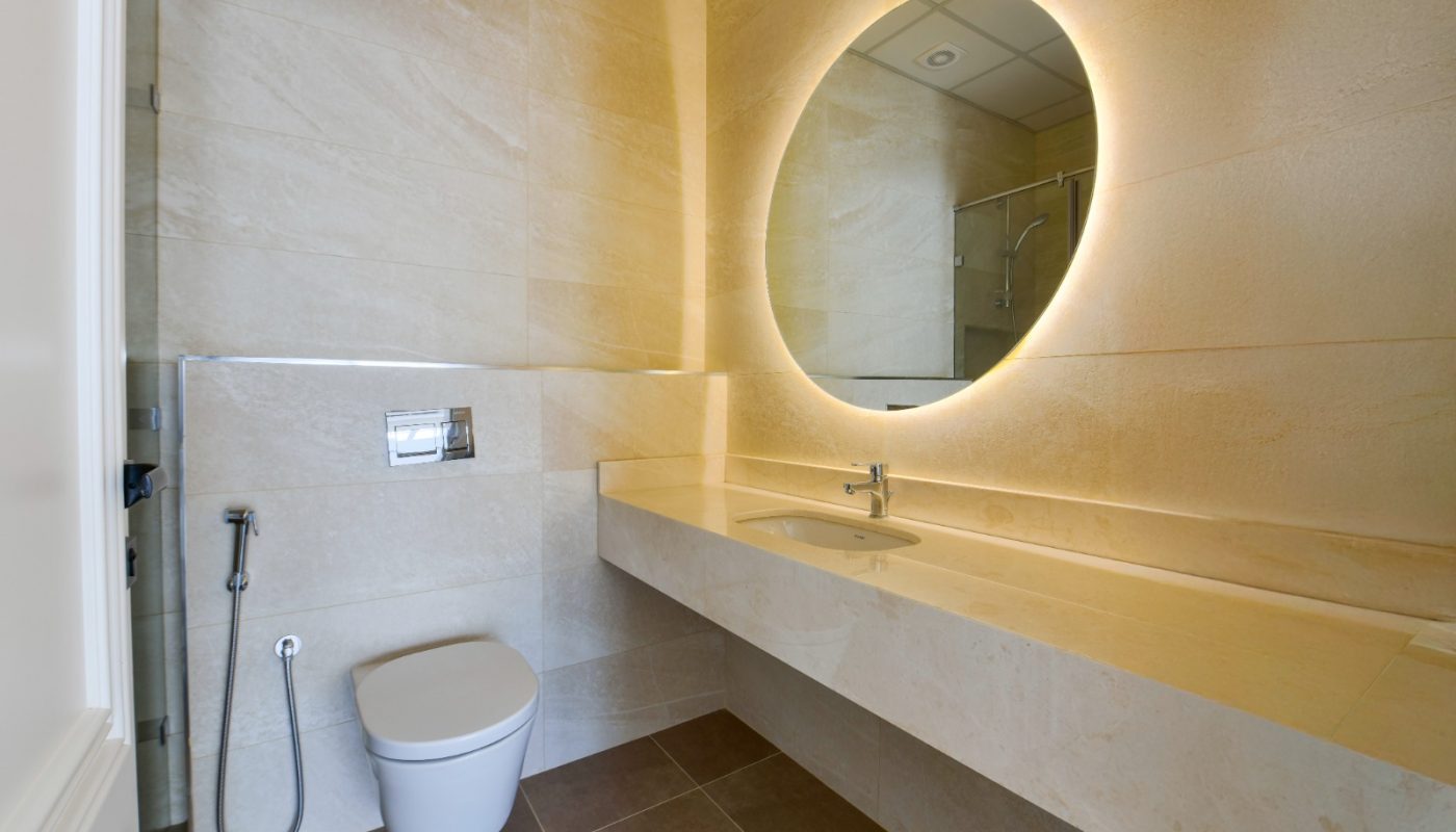 Modern bathroom with beige tiled walls, wall-mounted toilet, bidet spray, large countertop, and a round backlit mirror above the sink.
