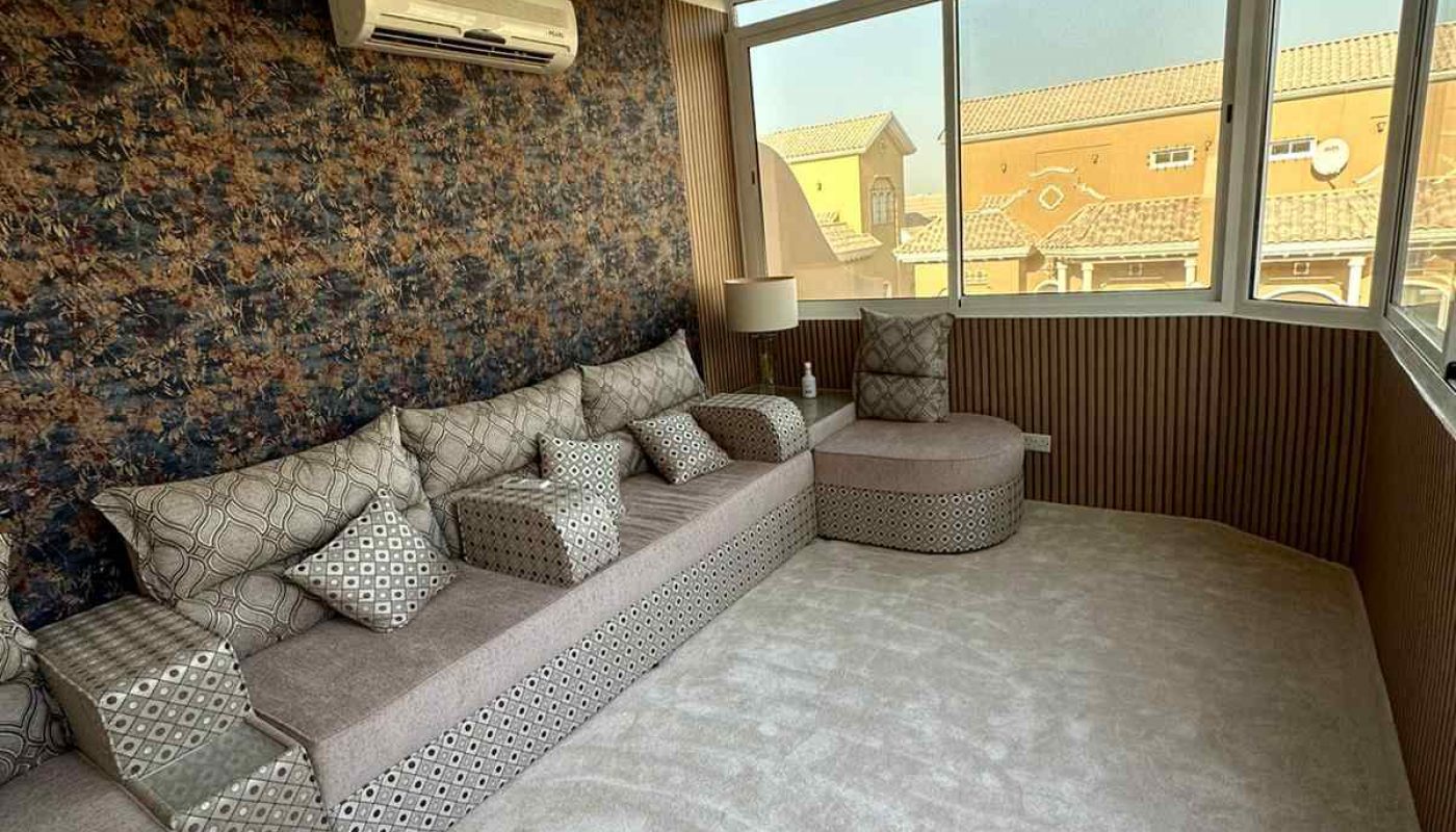 A sunlit room with patterned wallpaper, a long gray couch with matching pillows, a round ottoman, carpeted floor, and a split air conditioner beneath large windows.