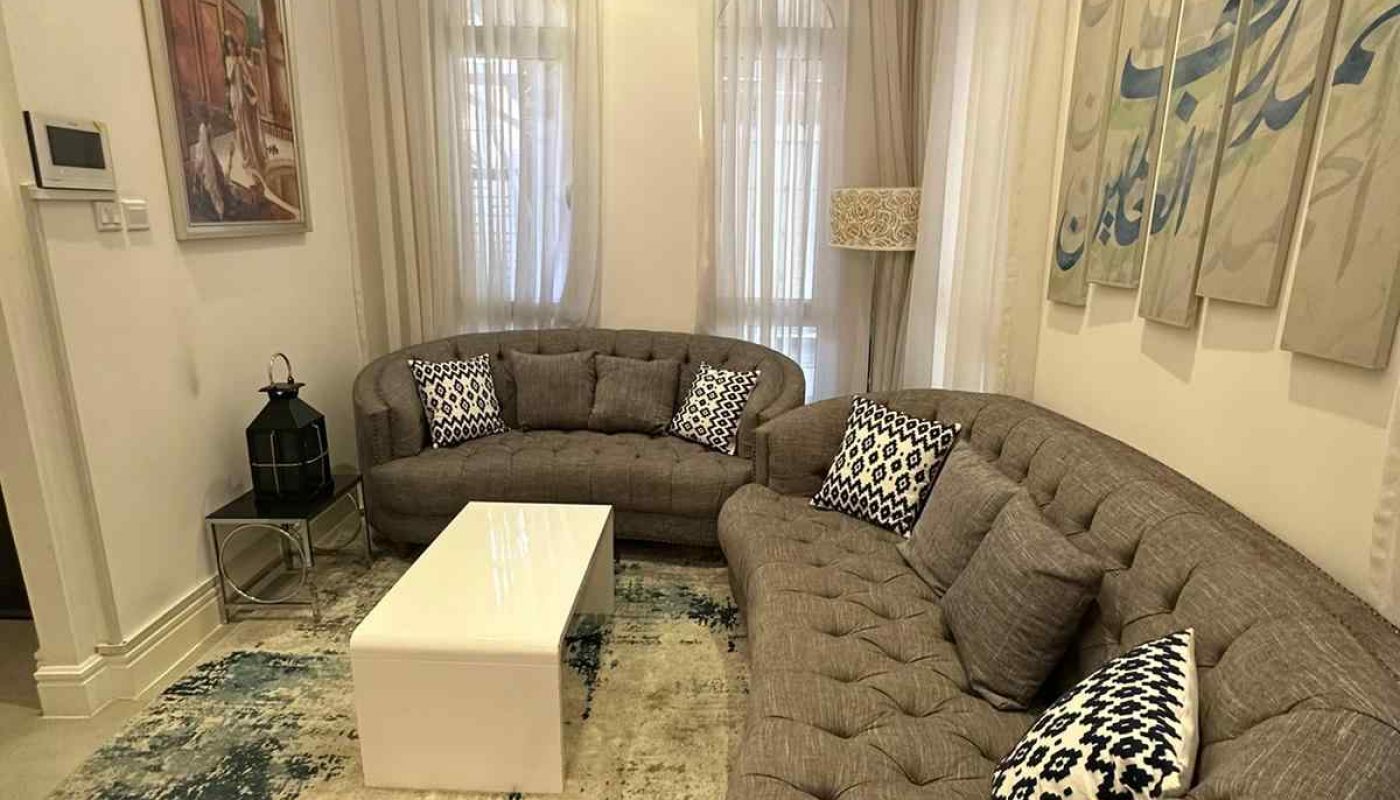 A living room with two curved gray sofas, a white rectangular coffee table, patterned rug, abstract wall art, and large windows with sheer curtains.