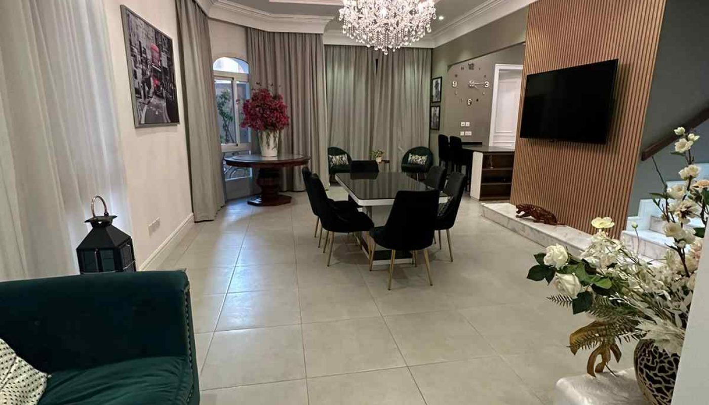 Modern living and dining area with a chandelier, green velvet sofa, dining table with six chairs, wall-mounted TV, large floor tiles, and decorative flowers.