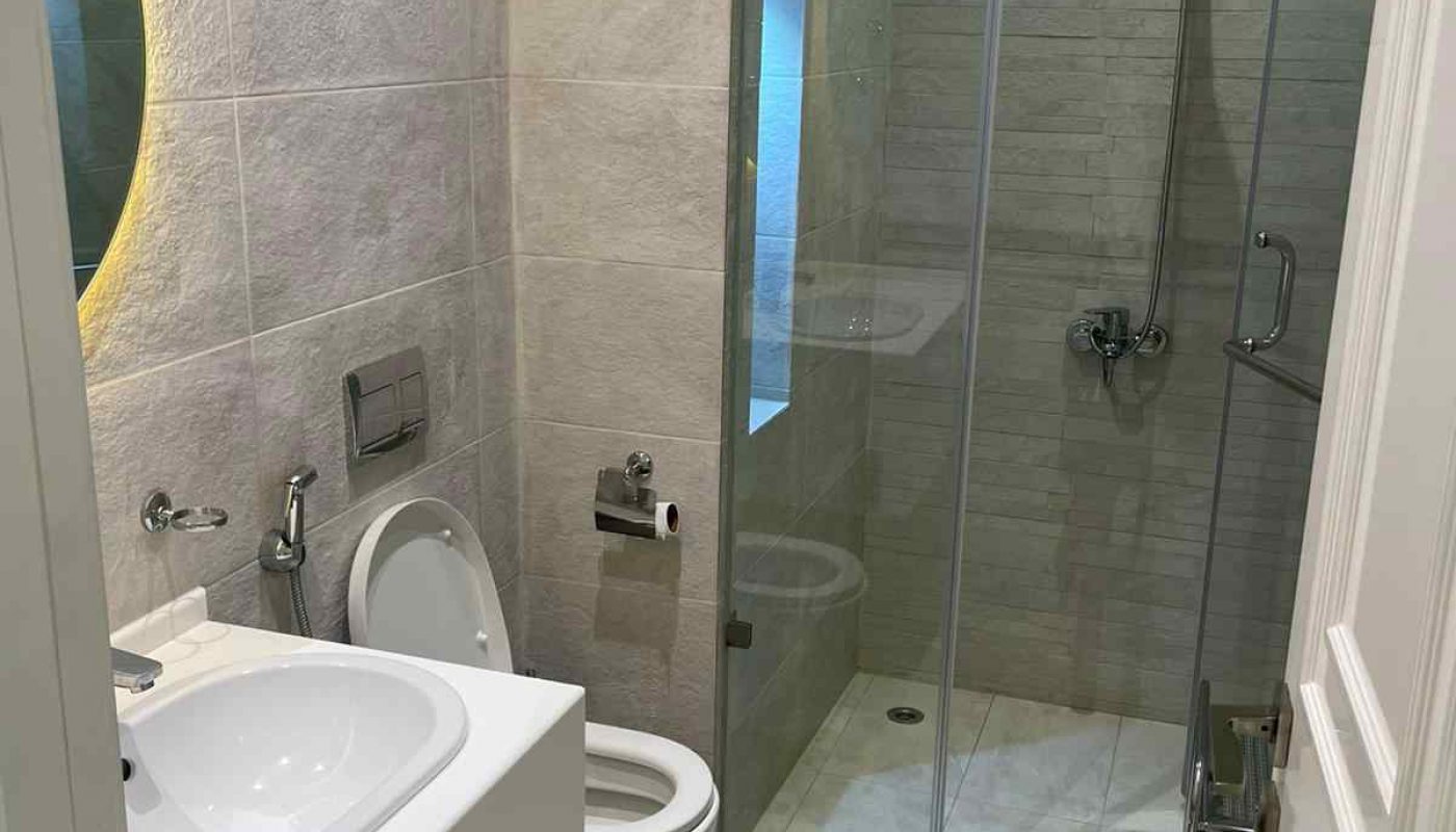 Modern bathroom with a white sink, wall-mounted toilet, glass-enclosed shower, and light gray tiled walls and floor.