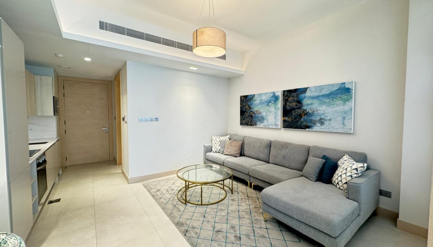 Modern living room with a grey sectional sofa, glass coffee table, abstract wall art, and an overhead light fixture. The space connects to a kitchen and has a neutral color palette.