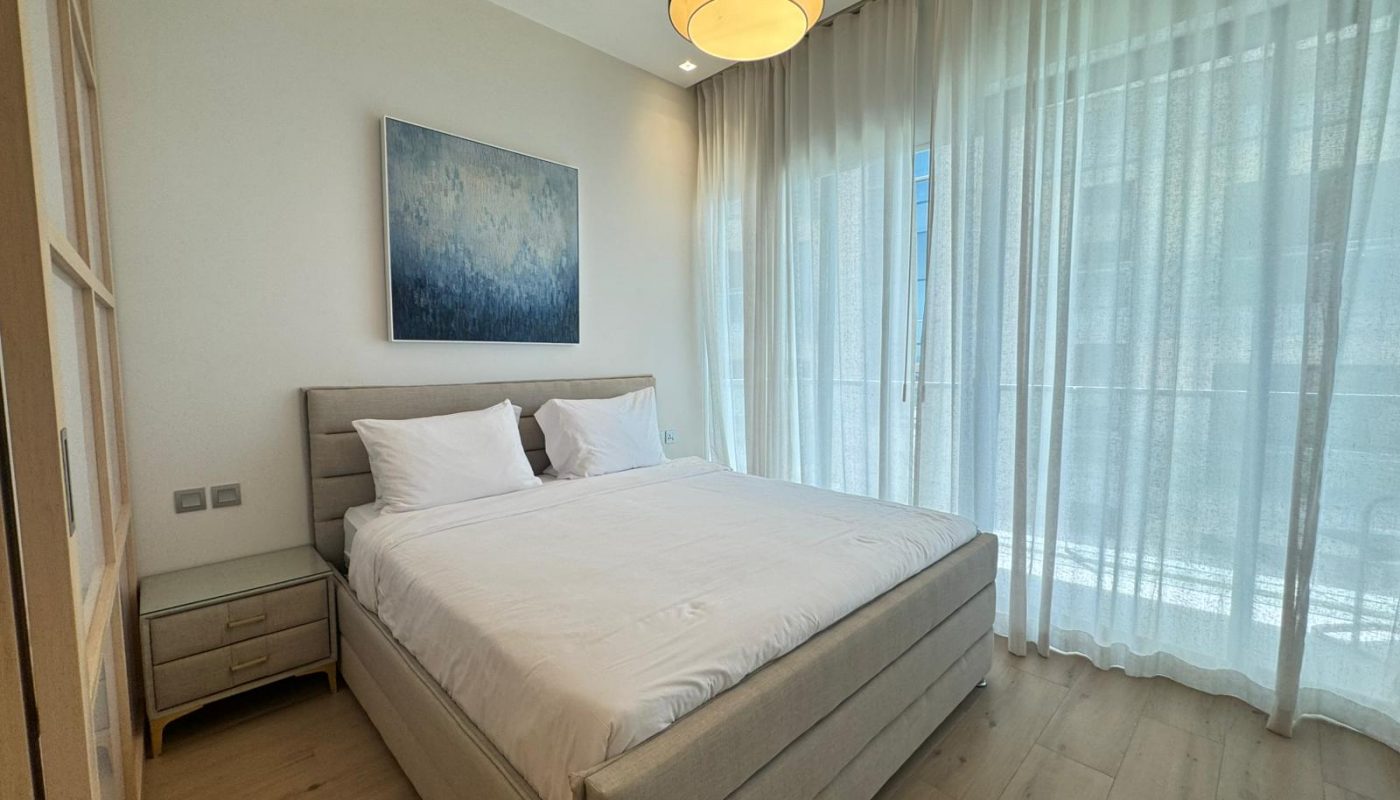 A modern bedroom with a double bed, white bedding, two pillows, a nightstand, sheer curtains, wood flooring, and a ceiling light fixture. A framed abstract artwork hangs on the wall.
