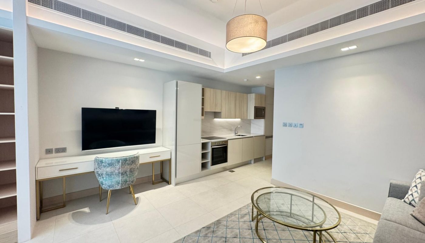 Modern living room and kitchen area with a TV on a desk, upholstered chair, round glass coffee table, gray sofa, and built-in shelves, featuring neutral decor and overhead lighting.