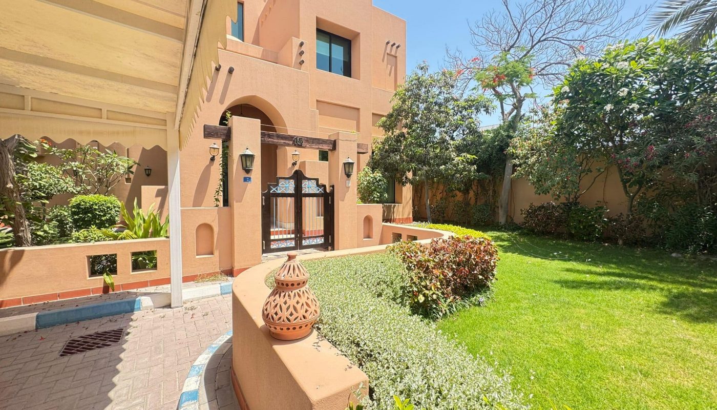 Sunlit garden with green lawn and shrubs in front of a modern two-story beige house, featuring a decorative gate and terracotta lantern on a low wall.
