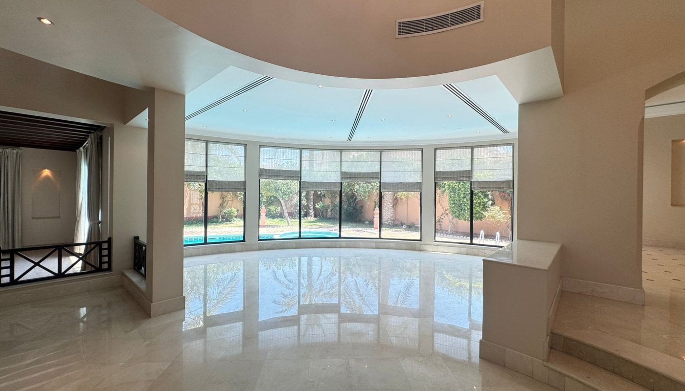 Spacious, light-filled room with large curved windows overlooking a swimming pool and garden area; marble flooring and neutral walls.