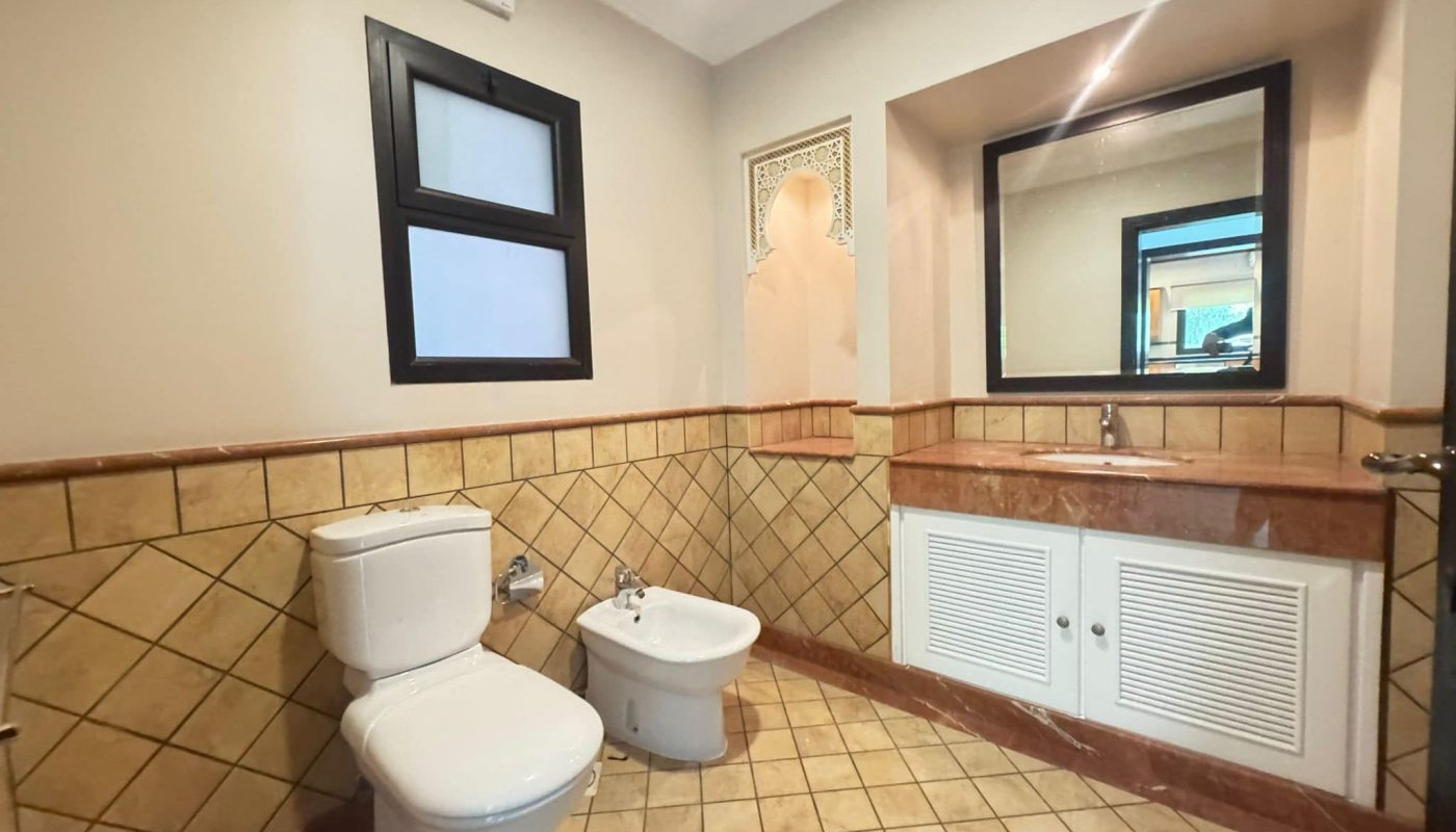 Bathroom with beige tiled floor and walls, featuring a toilet, bidet, large mirror, sink with cabinet, and a small frosted window.