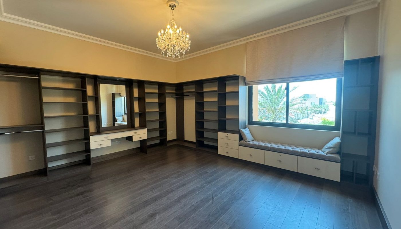Spacious walk-in closet with dark wood flooring, open shelving, a dressing table with drawers, a window seat, and a large window letting in natural light.