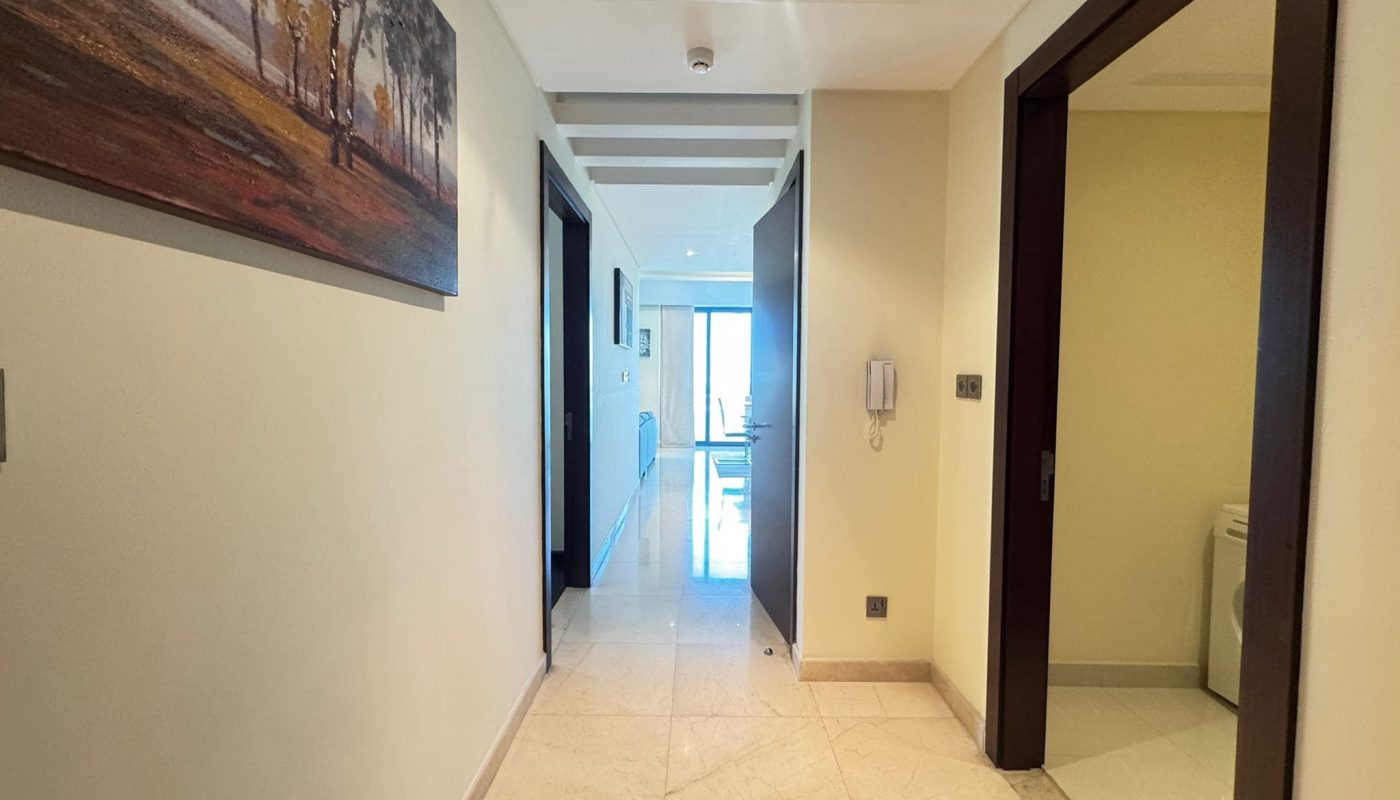 A hallway with beige tile flooring, doors on both sides, a wall-mounted painting on the left, and a view into a bright living area with large windows at the end.