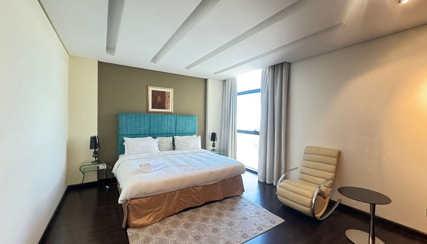 A modern bedroom with a double bed, turquoise headboard, side tables with lamps, a beige lounge chair, a round table, and a large window with curtains.