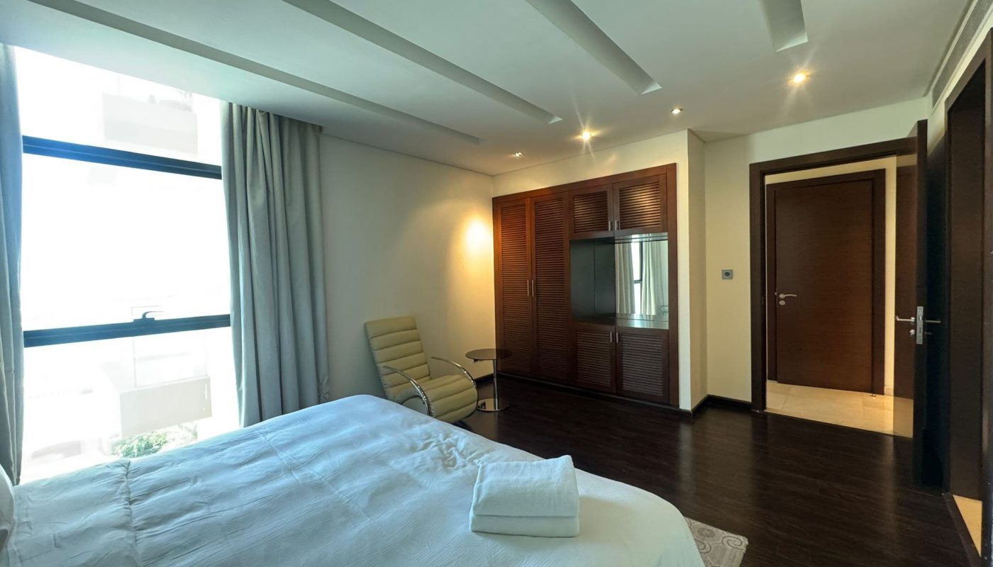 Modern bedroom with a neatly made bed, armchair, wardrobe with TV, large window with curtains, and dark wood flooring. Towels are placed on the bed.