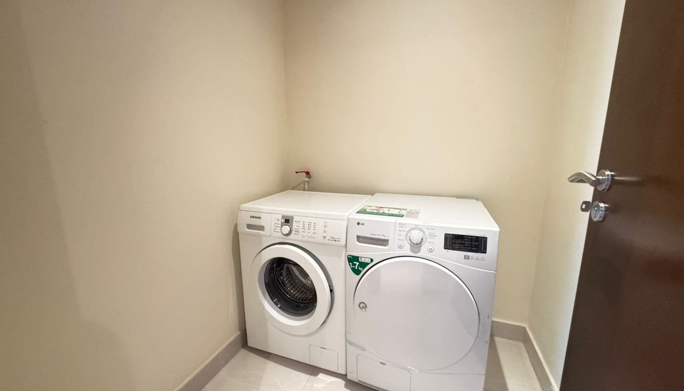 A white washing machine and dryer are placed side by side in the corner of a small, empty laundry room with beige walls and tiled floor.