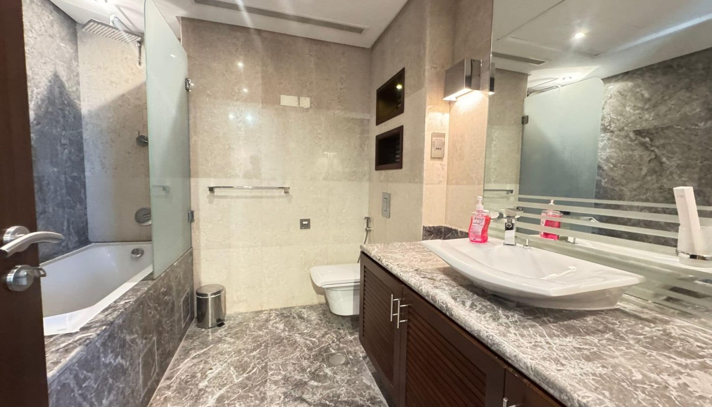 Modern bathroom with marble surfaces, a wall-mounted toilet, a bathtub with a glass partition, a large mirror, and a countertop with a vessel sink and toiletries.