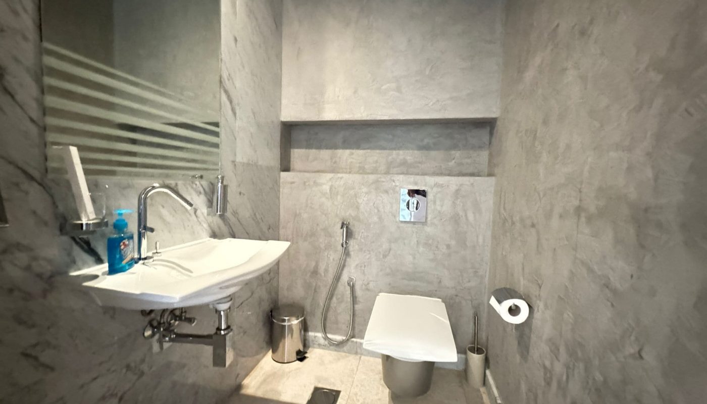 A modern bathroom with grey walls, a wall-mounted sink and faucet, a toilet with a bidet spray, a trash bin, and a mirror on the left side.