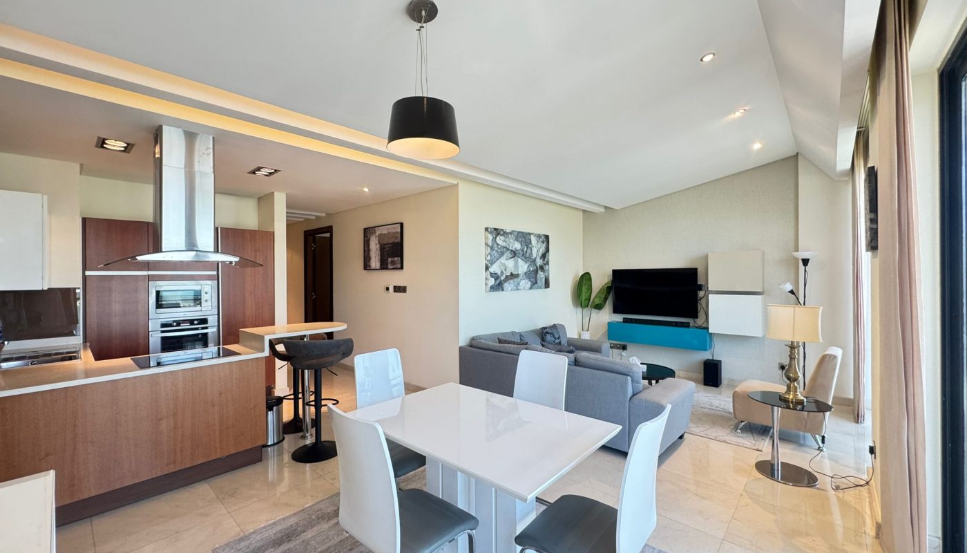 Modern open-plan living space with a kitchen, dining table, and living area featuring neutral tones, contemporary furniture, and large windows letting in natural light.