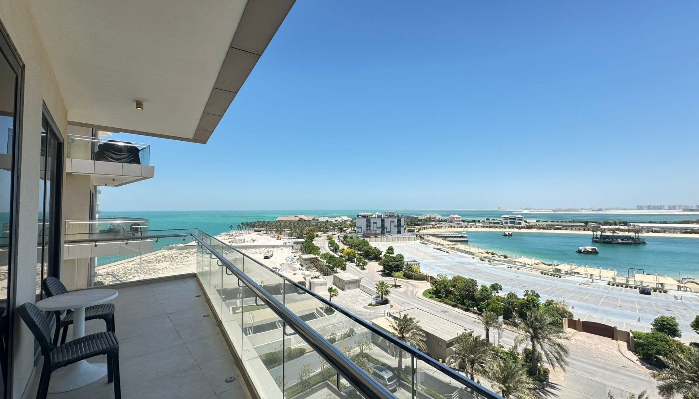 A modern balcony with chairs overlooks a road, palm trees, buildings, and a beach with blue sea under a clear sky.