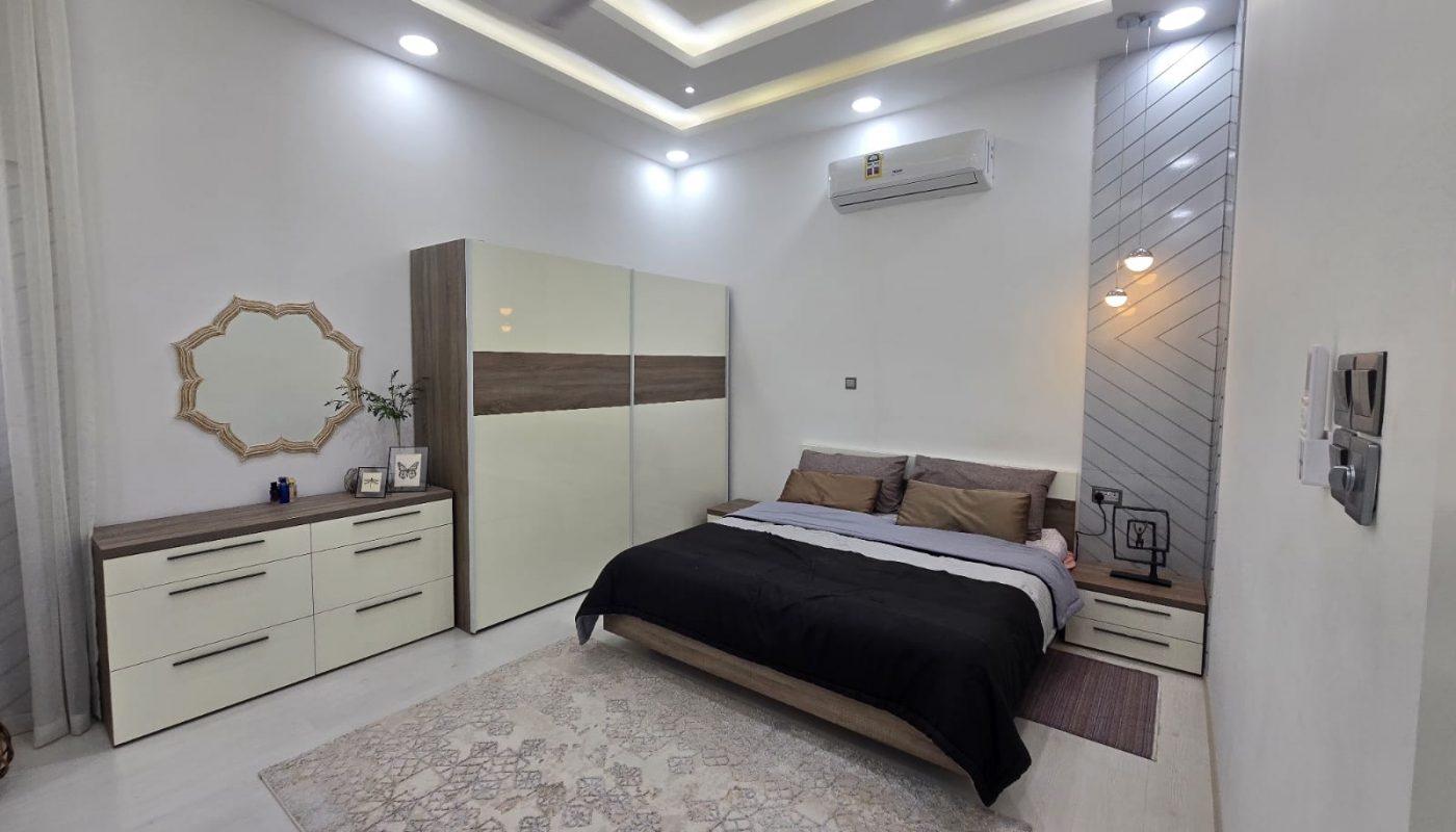 Modern bedroom with a double bed, beige and brown bedding, a large wardrobe, dresser with mirror, area rug, ceiling fan, air conditioner, and geometric accent wall with pendant light.