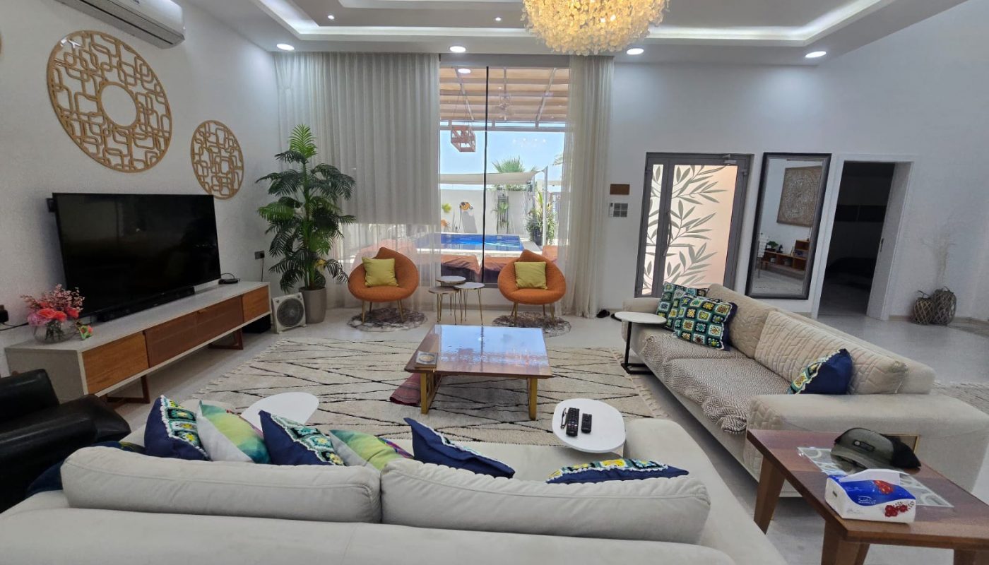 Modern living room with two beige sofas, colorful pillows, a TV, two orange chairs, a coffee table, and large windows overlooking a pool. Decorative wall art and a chandelier are present.